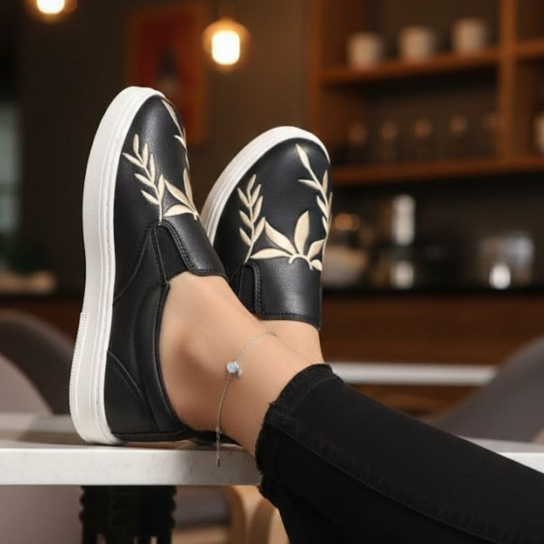 Person wearing black slip-on shoes with white soles and leaf patterns, sitting on a table indoors.