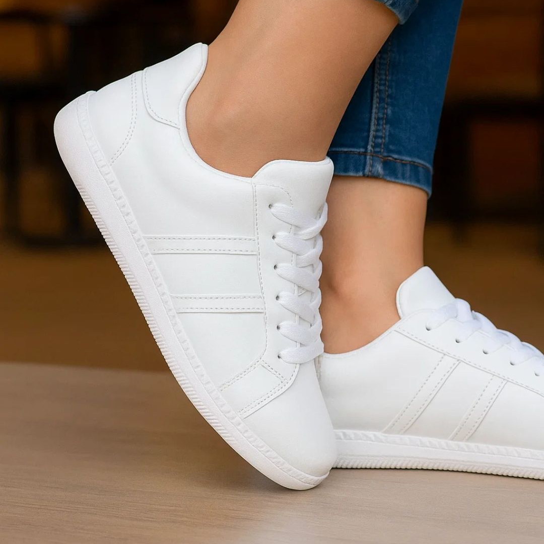 White sneakers worn with blue jeans on a wooden floor.