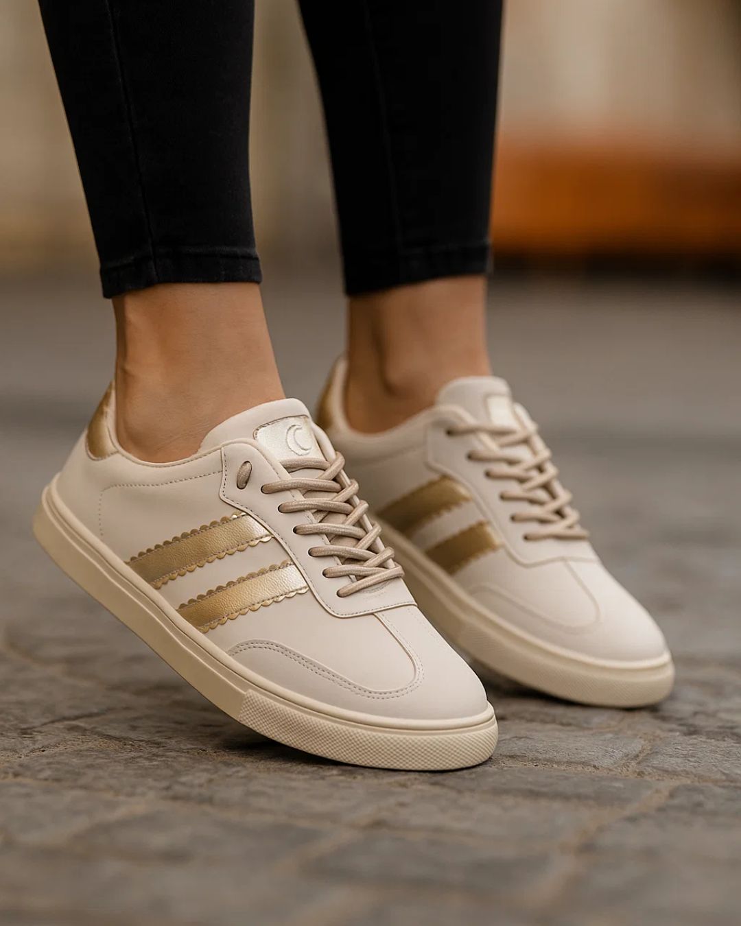 White sneakers with gold stripes worn by a person on a stone floor.