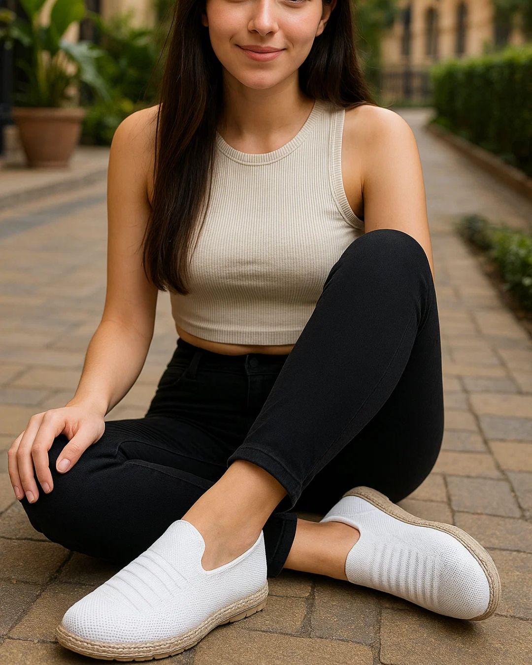 Woman sitting outdoors wearing a beige tank top, black pants, and white slip-on shoes.