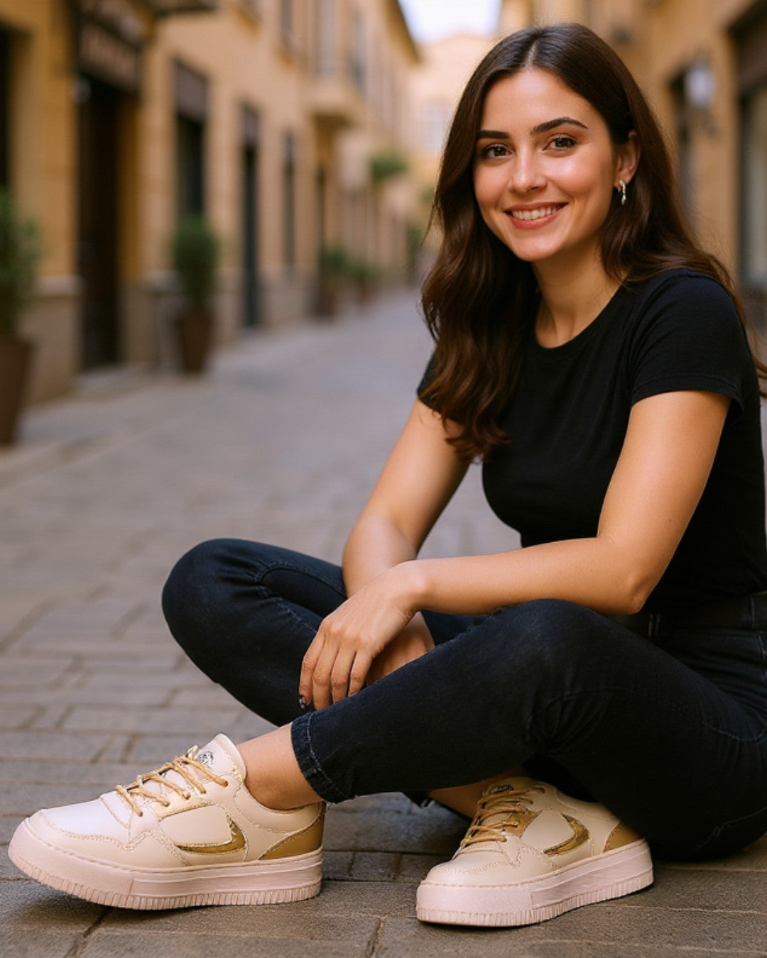 Woman sitting on a street wearing black t-shirt, dark jeans, and cream sneakers with gold accents.