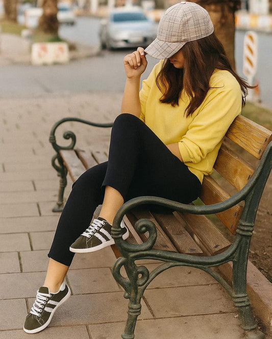 Woman sitting on a bench wearing a yellow sweatshirt and plaid cap in an urban setting.
