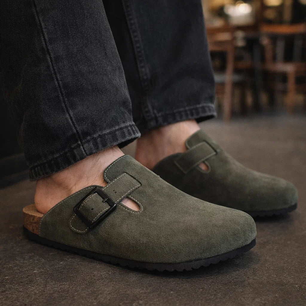 Green suede clogs worn with dark jeans on a blurred indoor background