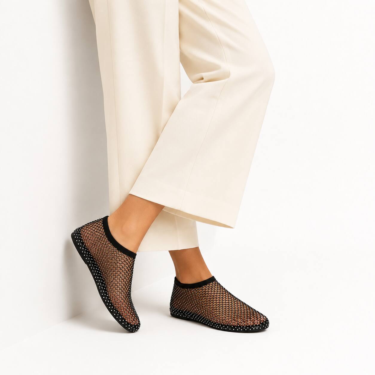 A person wearing black mesh slip-on socks with black trim, paired with cream wide-leg trousers, standing against a white wall.
