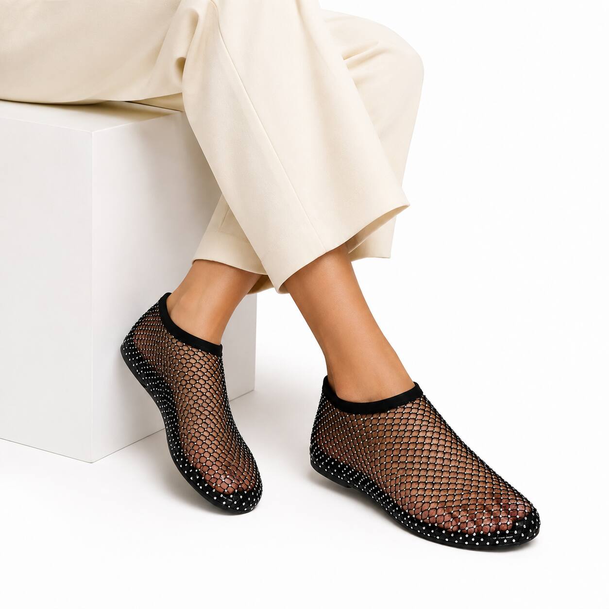 A person wearing black mesh slip-on rain boots with black soles and small silver studs, paired with cream pants, sitting on a white block against a white background.