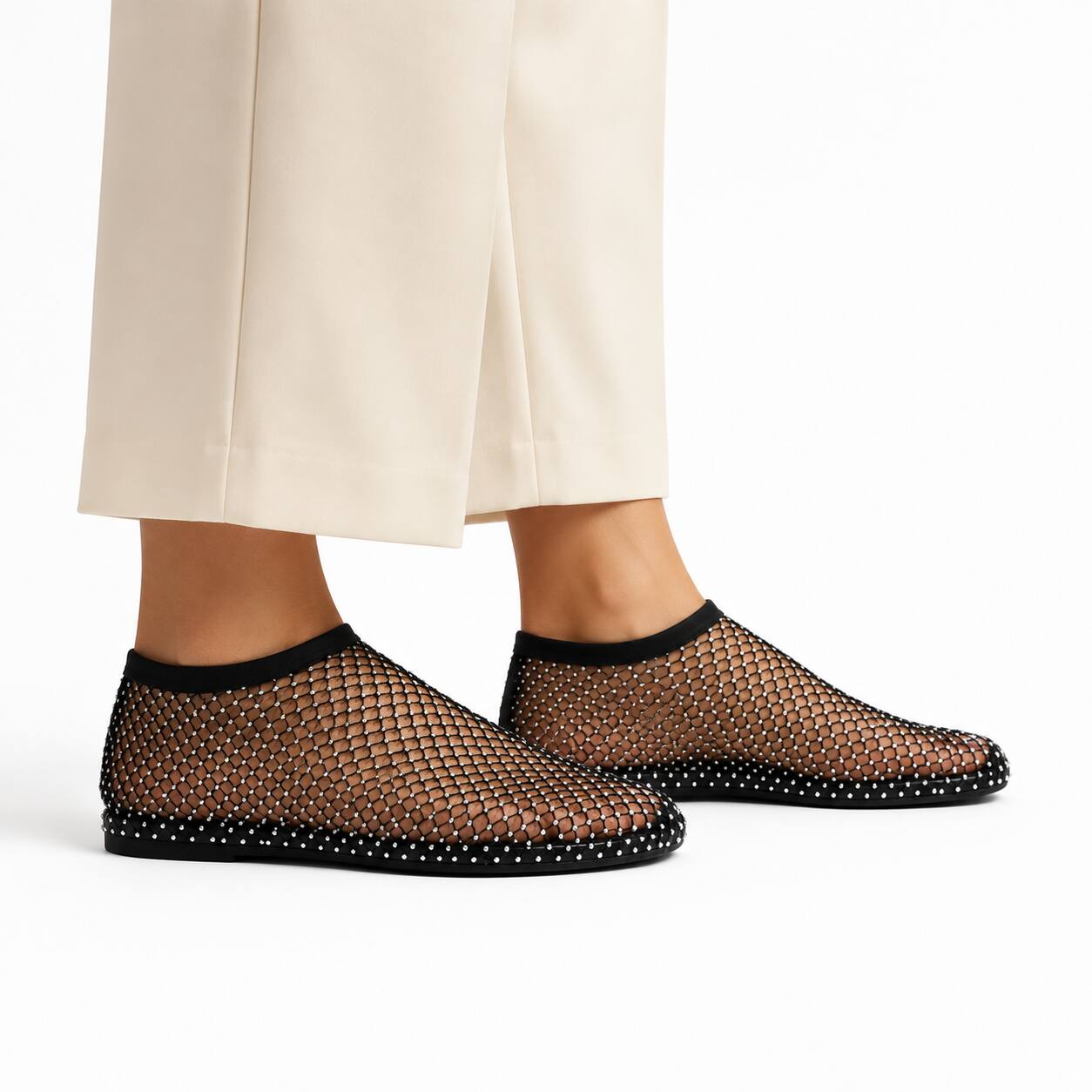 A person wearing beige cropped trousers and black mesh slip-on shoes with a studded black sole, shown against a clean white studio background.