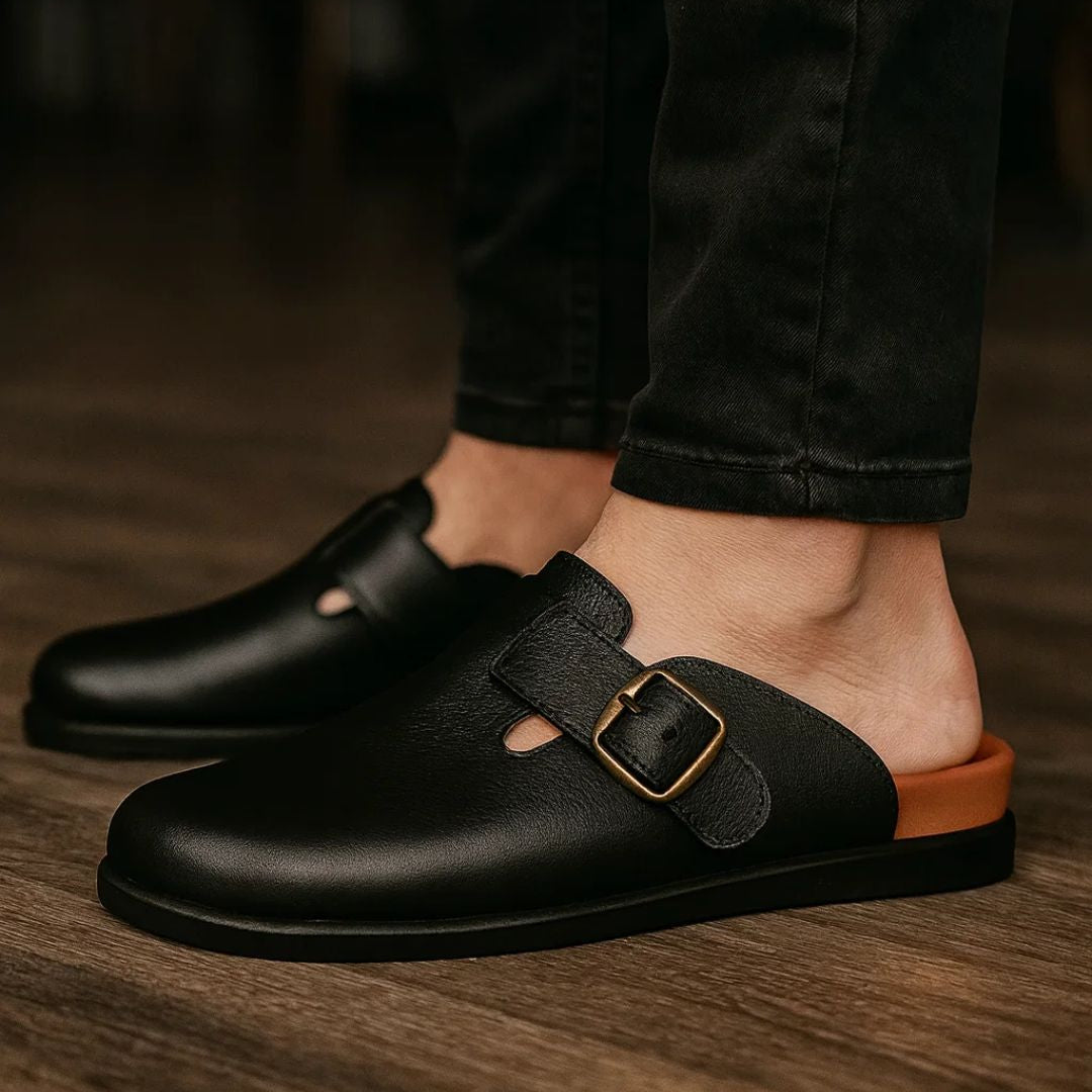 Black leather shoes with a brown sole worn by a person on a wooden floor.