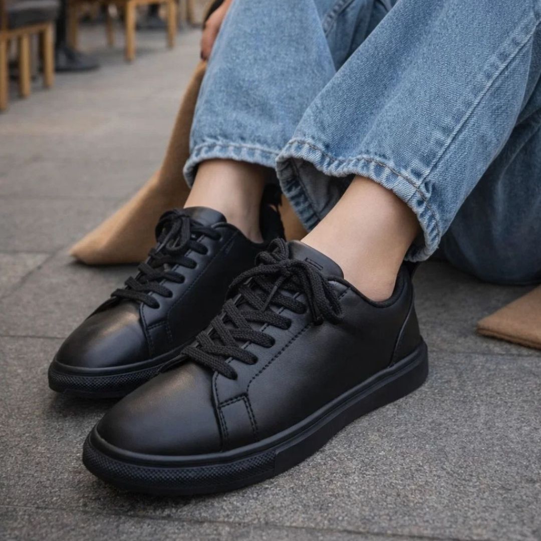 Black leather shoes worn with blue jeans on a pavement.
