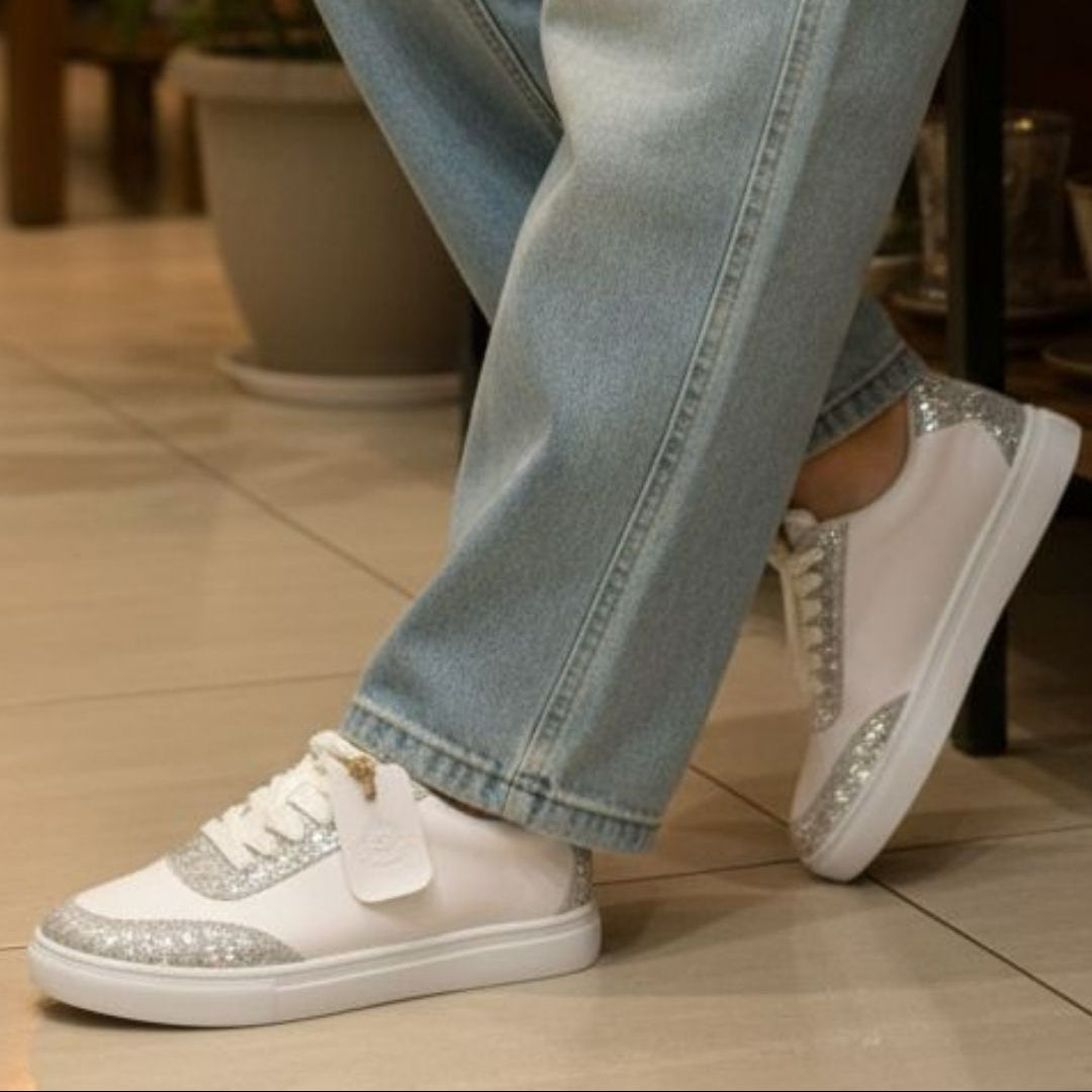 White sneakers with glittery details worn with light blue jeans on a tiled floor.