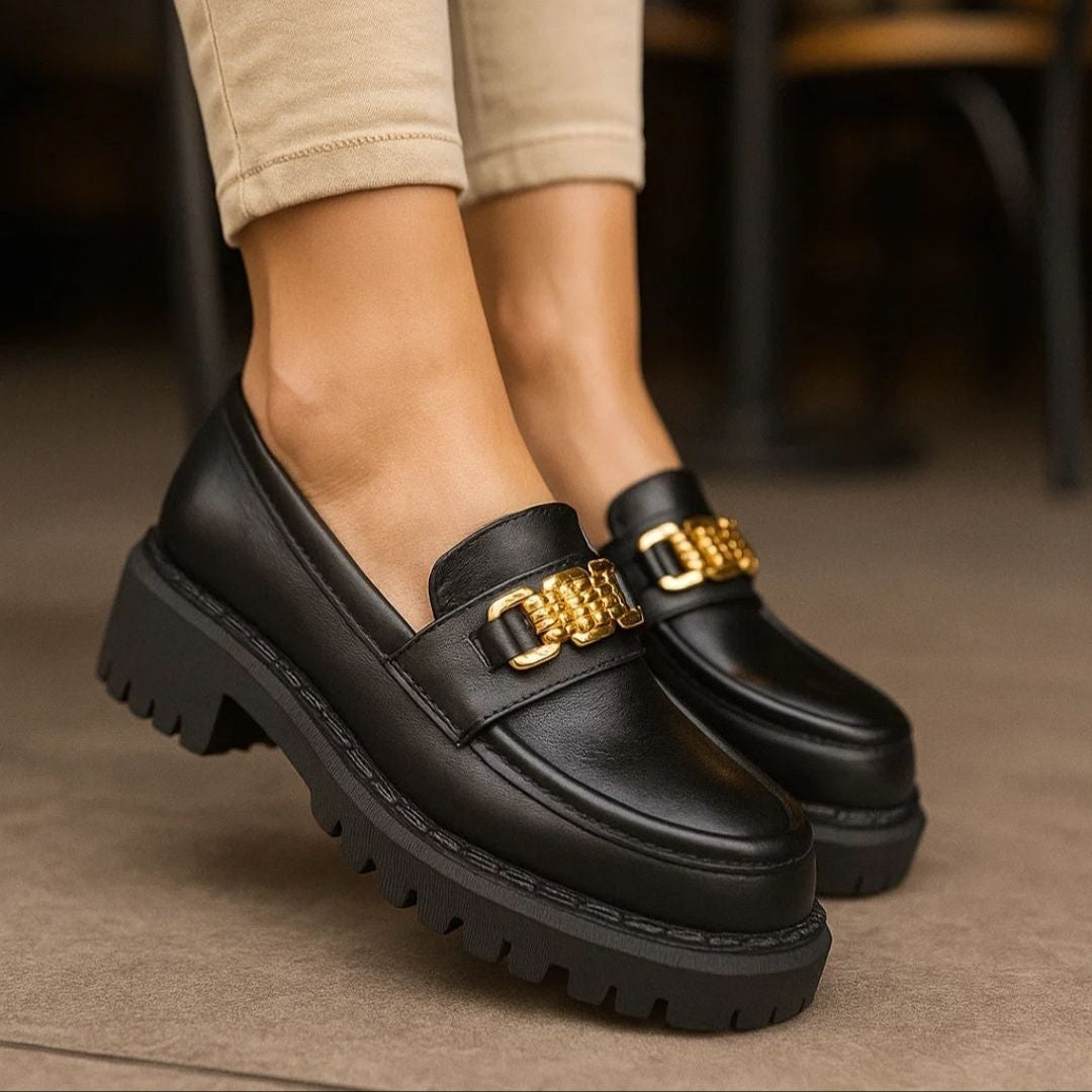 Black loafers with gold chain details worn by a person on a neutral background