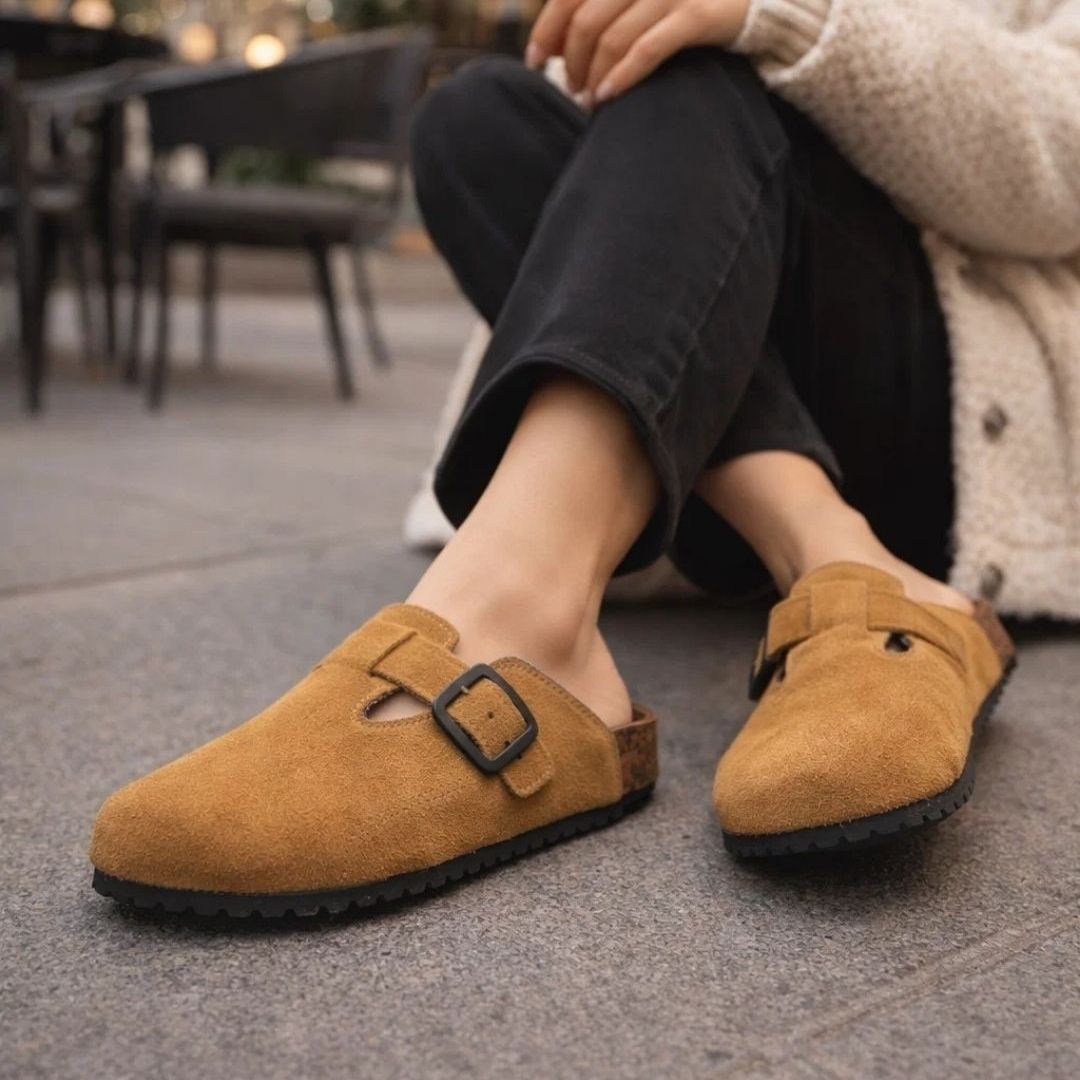 Brown suede clogs worn by a person sitting outdoors.