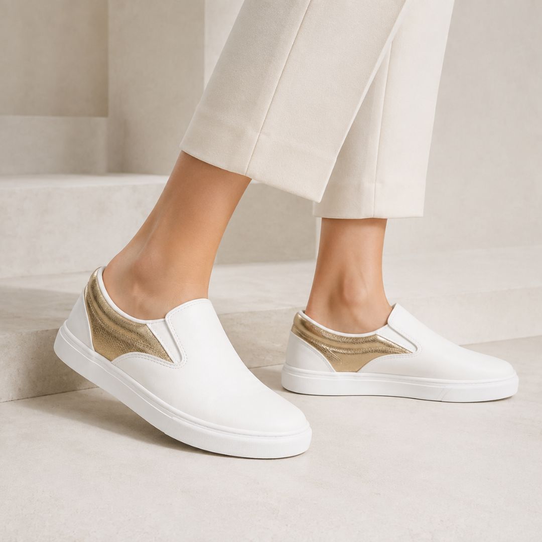 White slip-on sneakers with metallic gold wedge accents on the sides, shown on a model wearing off-white cropped trousers against a minimalist beige stone staircase background.