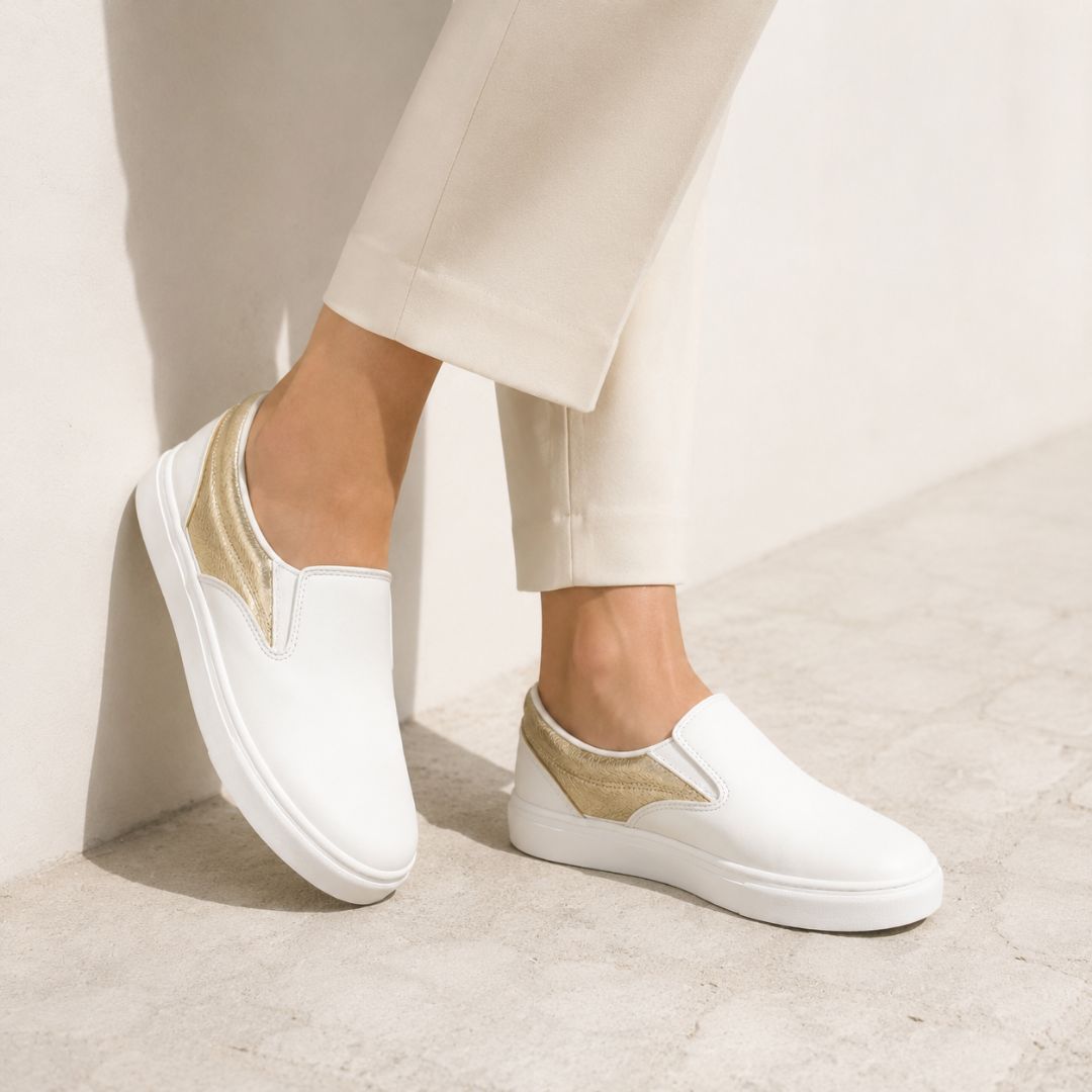 White slip-on platform sneakers with metallic gold side panels, shown on a model wearing beige trousers in a minimalist indoor setting.
