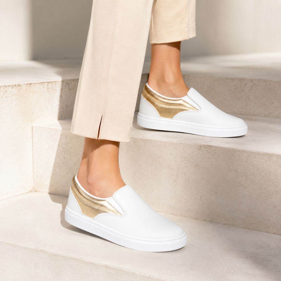 White slip-on sneakers with metallic gold side accents, shown on a model wearing beige trousers while standing on light stone steps.
