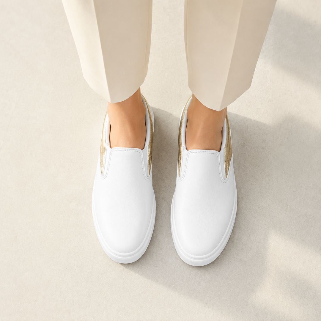 White slip-on sneakers with metallic gold side accents, shown on a model wearing beige trousers against a light beige background.