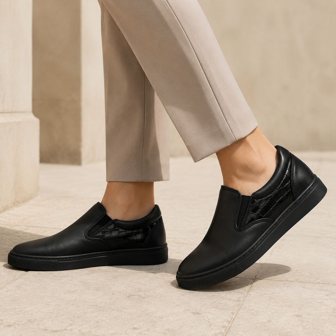 A person wearing black slip-on loafers with textured side panels, paired with beige trousers, standing on a light stone floor.