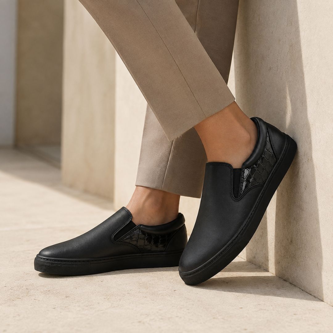 A person wearing black slip-on shoes and beige trousers, leaning against a light stone wall in a sunlit architectural setting.