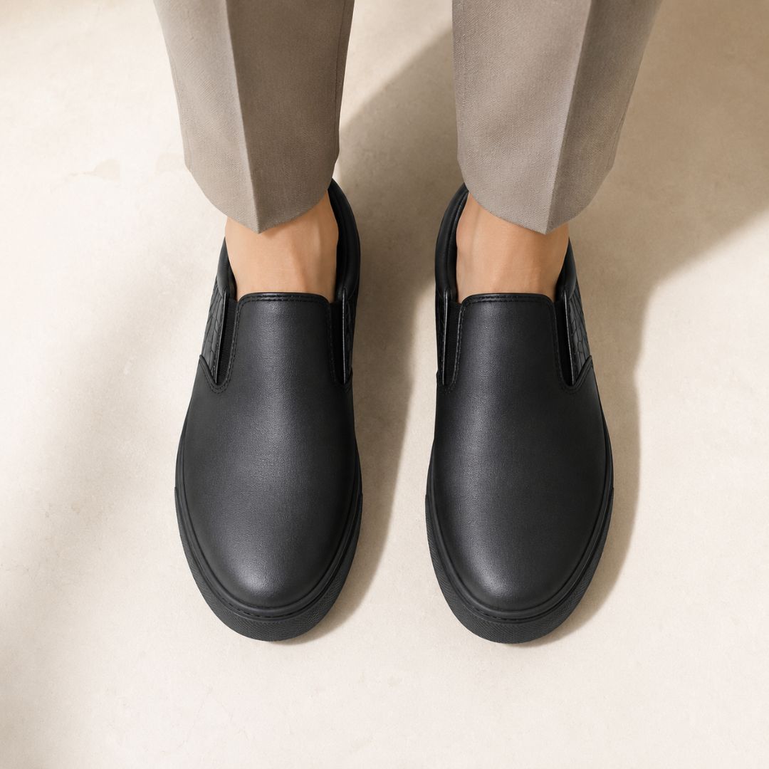 A pair of black slip-on shoes worn with light beige trousers, shown from a top-down view on a neutral beige floor.