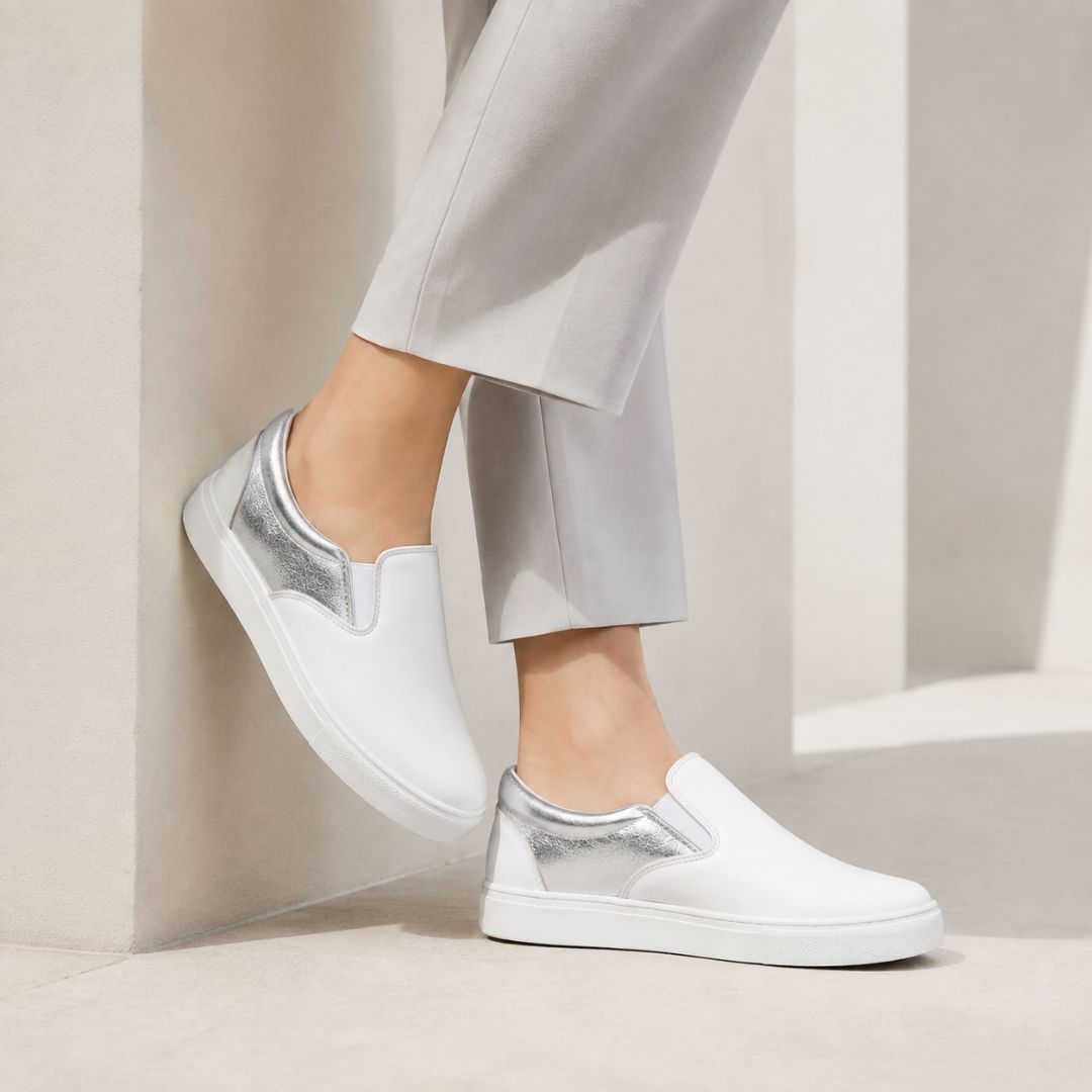 White slip-on sneakers with metallic silver heel panels, shown on a model wearing light gray trousers in a minimalist beige architectural setting.