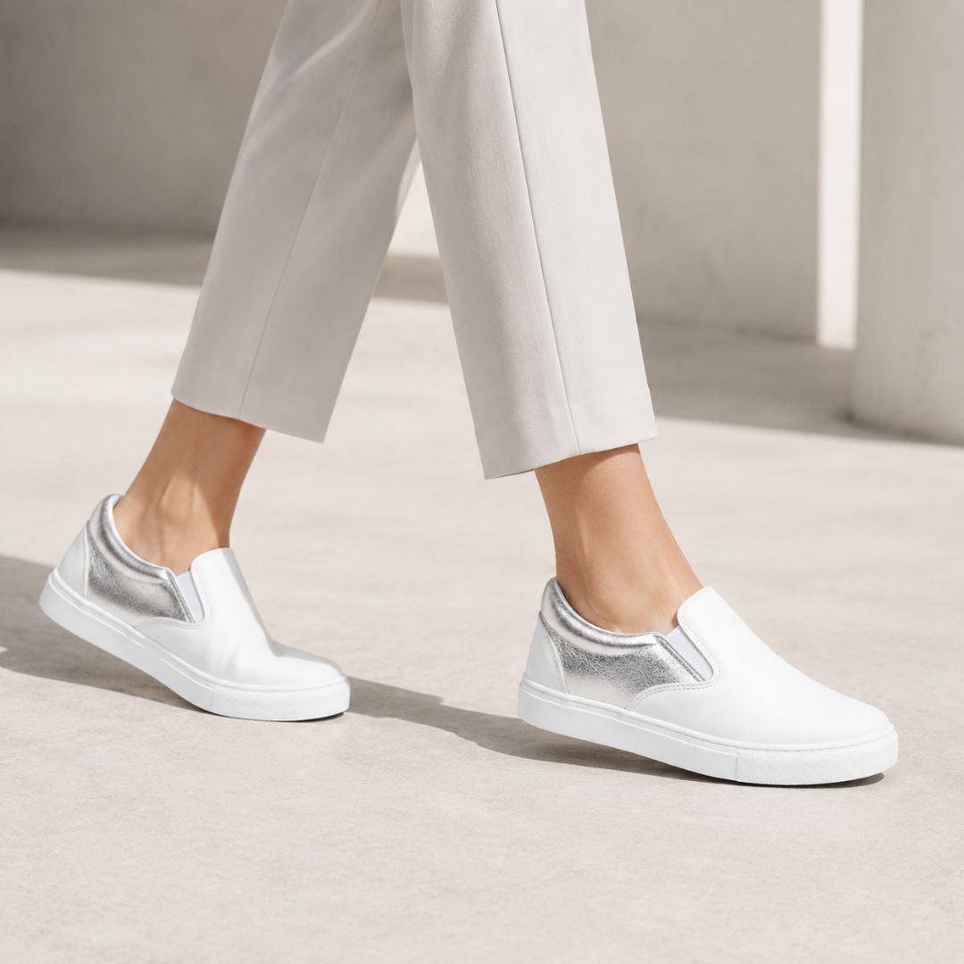 White slip-on sneakers with metallic silver side panels, shown on a model wearing light beige trousers in a minimalist concrete setting.