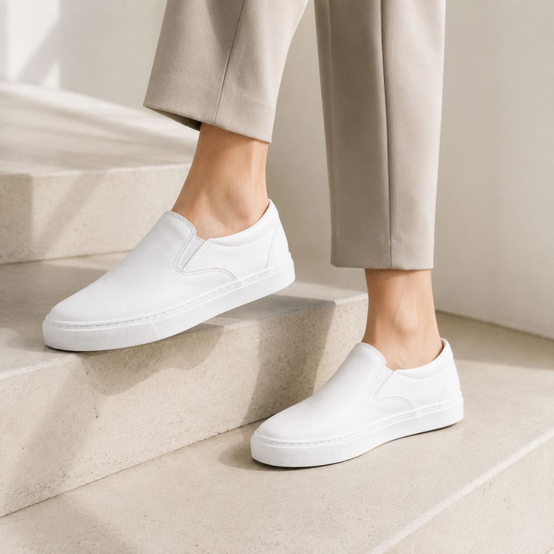 White slip-on canvas sneakers worn with beige trousers, shown on a person standing on light stone steps.