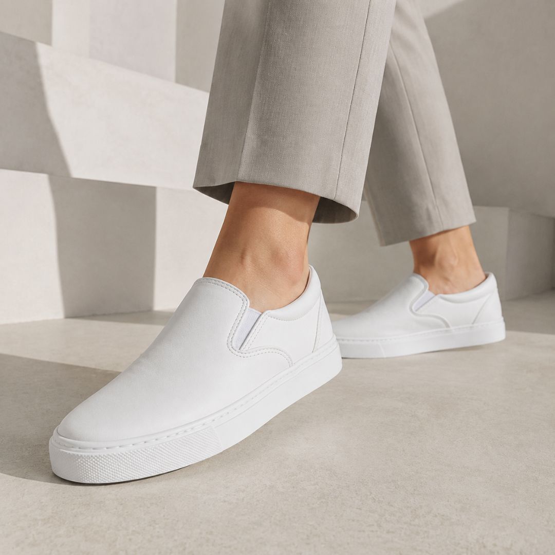 White slip-on platform sneakers worn with light gray trousers, shown on a model in a minimalist concrete setting.