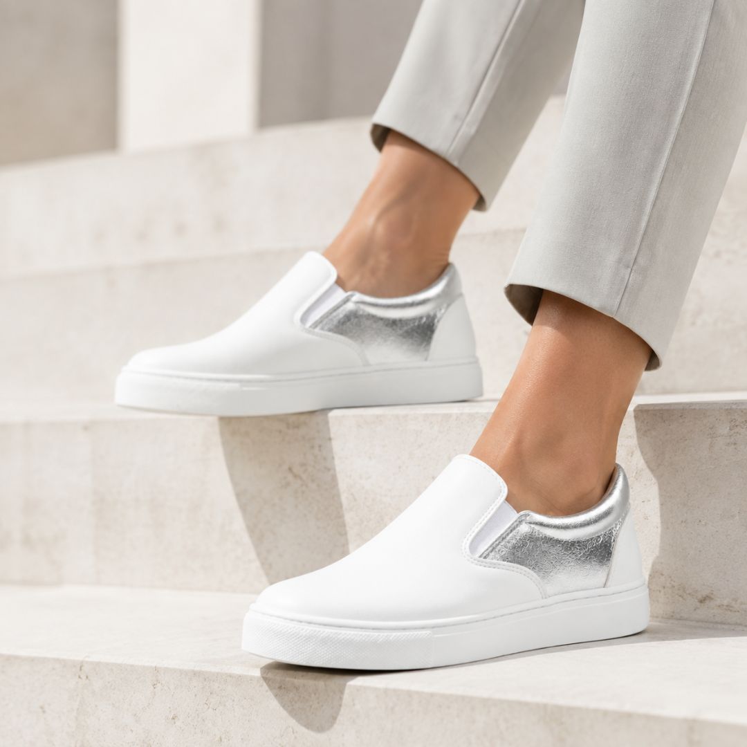 White slip-on sneakers with metallic silver heel accents, shown on a model wearing light gray trousers while sitting on light beige stone steps.
