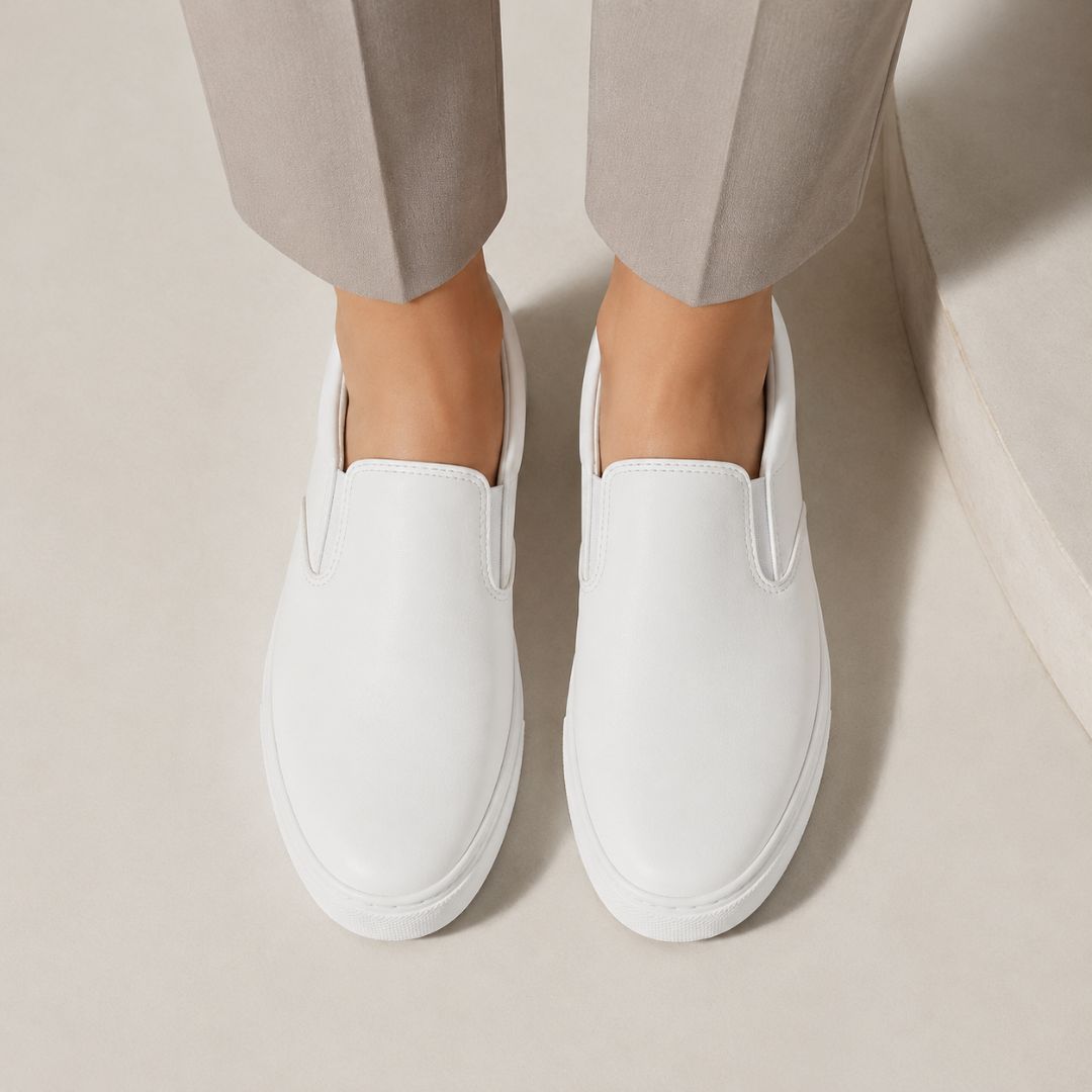 A pair of white slip-on leather shoes worn with light beige trousers, shown from a top-down angle on a neutral beige background.