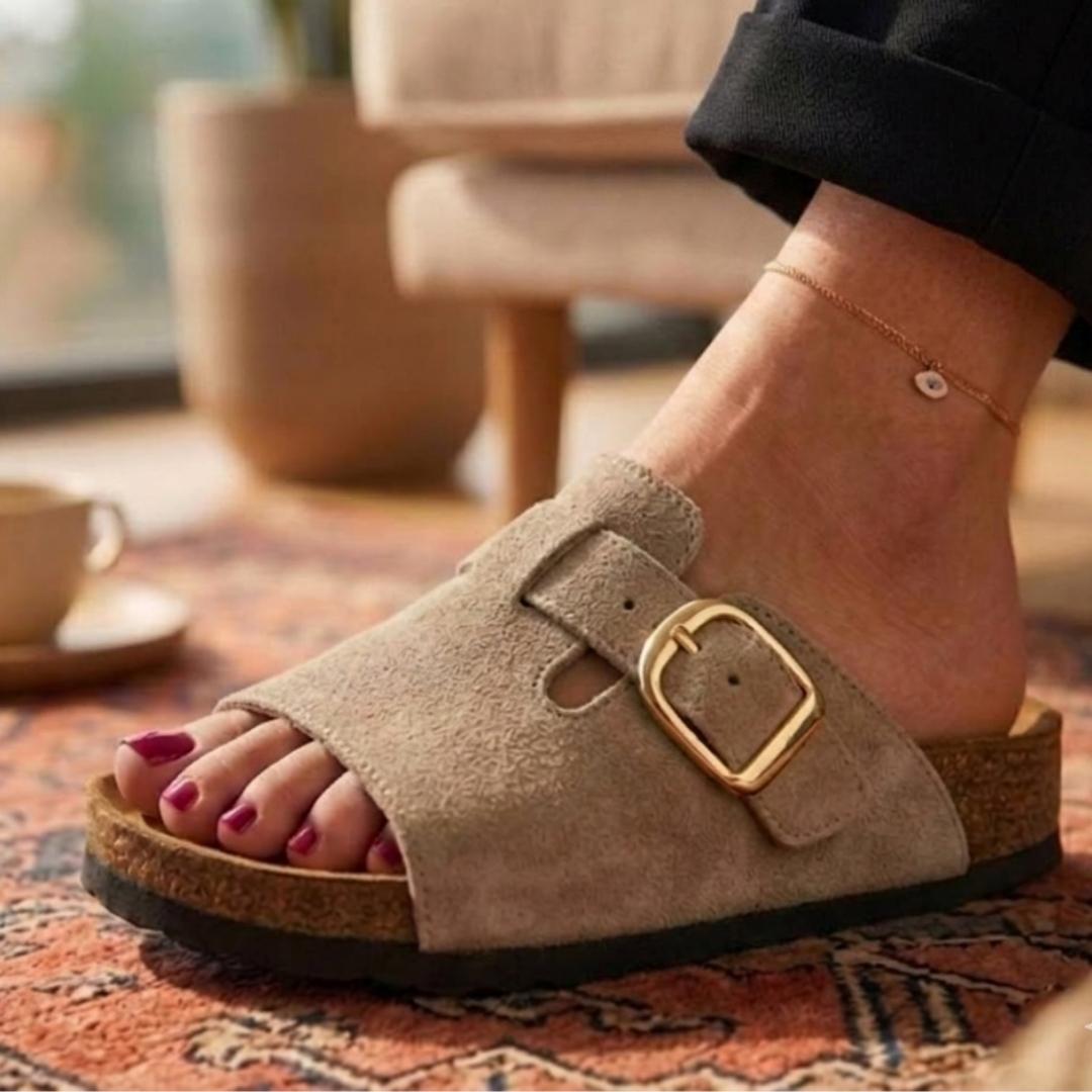 Brown suede sandal with gold buckle worn indoors on a patterned rug.