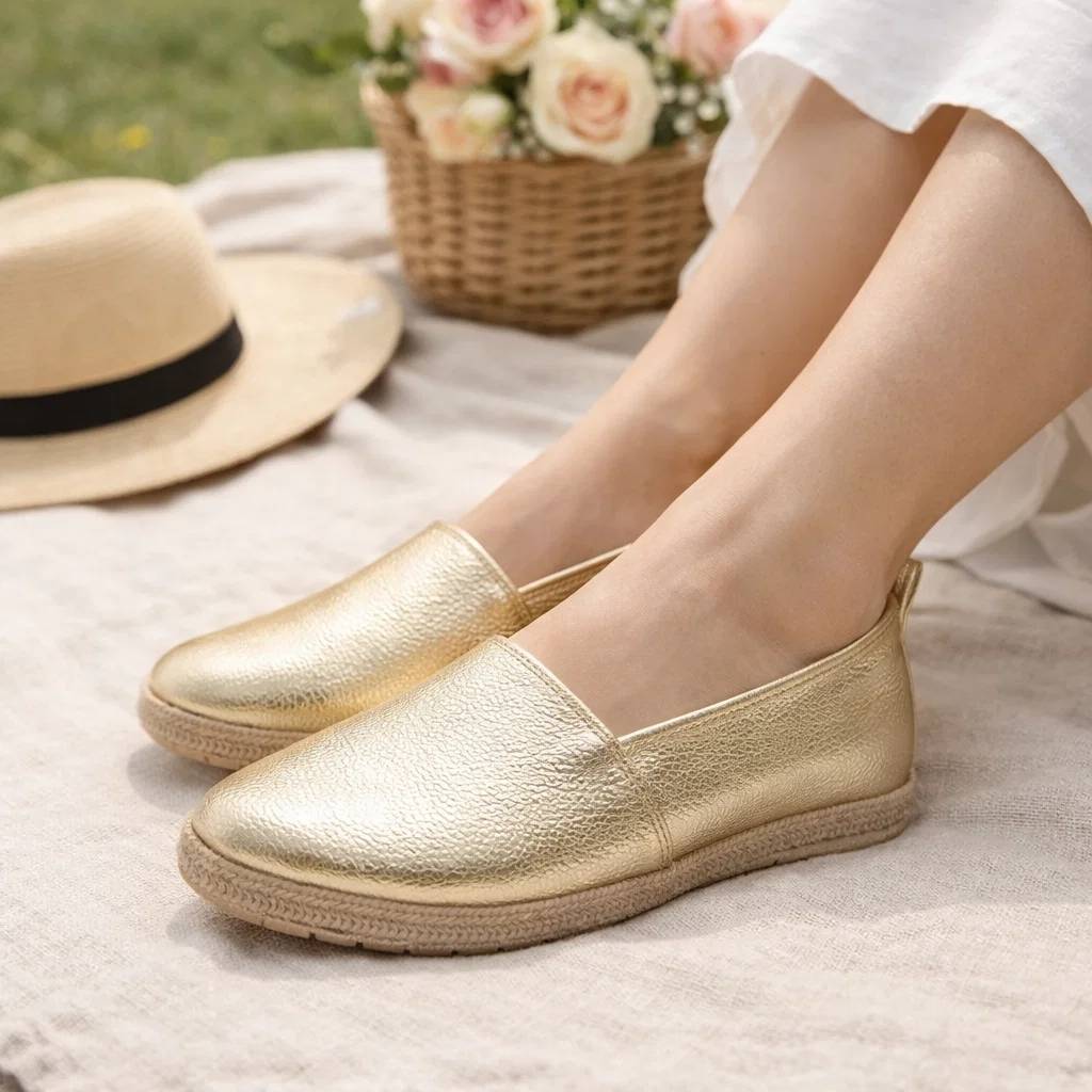 Gold slip-on shoes worn by a person sitting outdoors with a hat and flowers in the background.