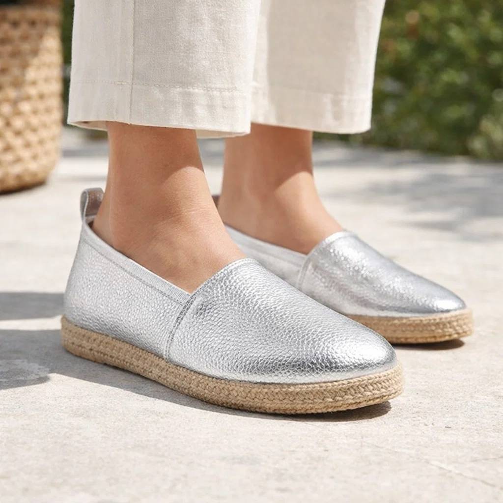 Silver espadrille shoes worn with light-colored pants on a blurred outdoor background