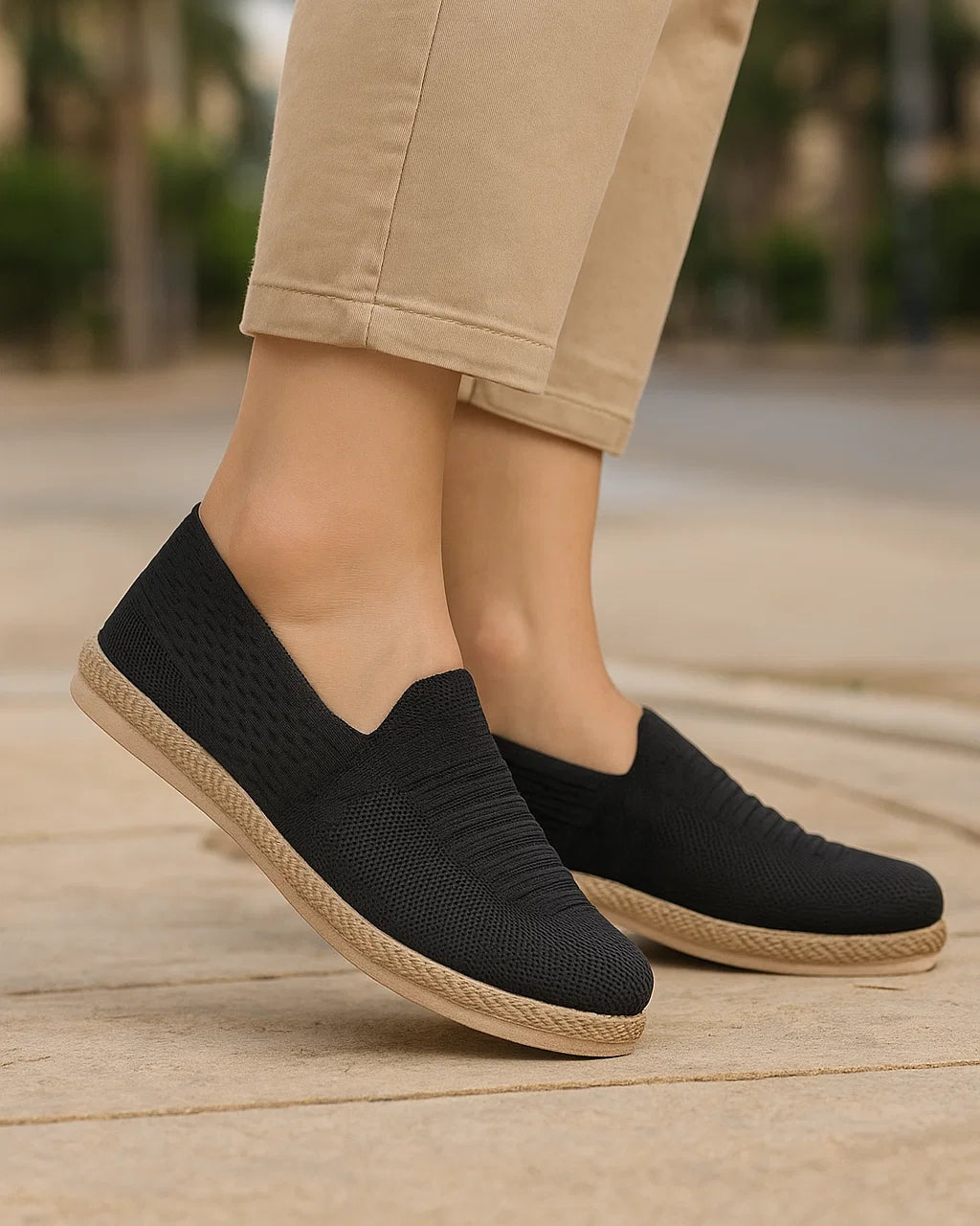 Black slip-on shoes with beige soles worn with beige pants on a wooden floor.