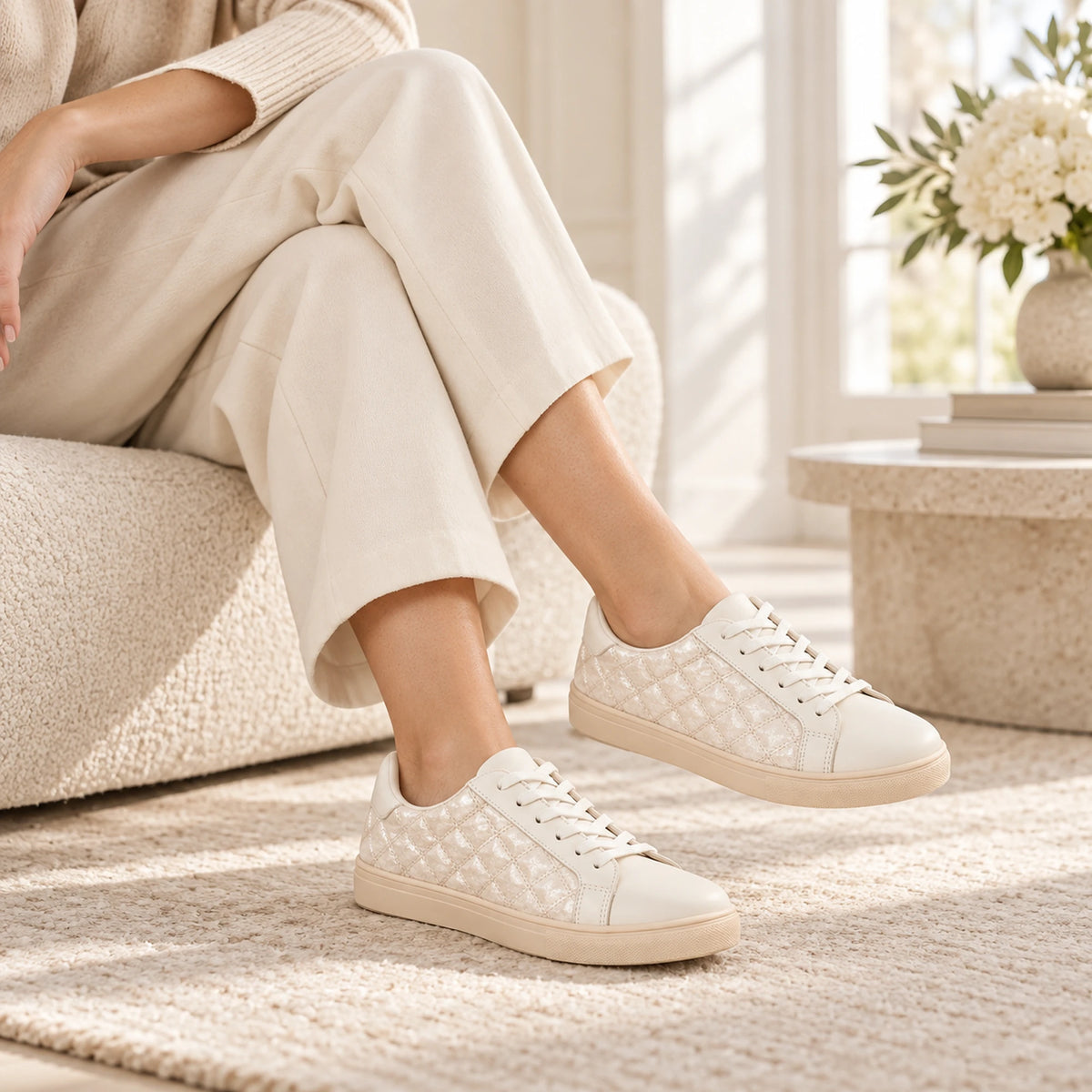 A person seated on a textured beige sofa wearing cream-colored low-top sneakers with a quilted geometric pattern on the sides, in a bright, sunlit living room setting.