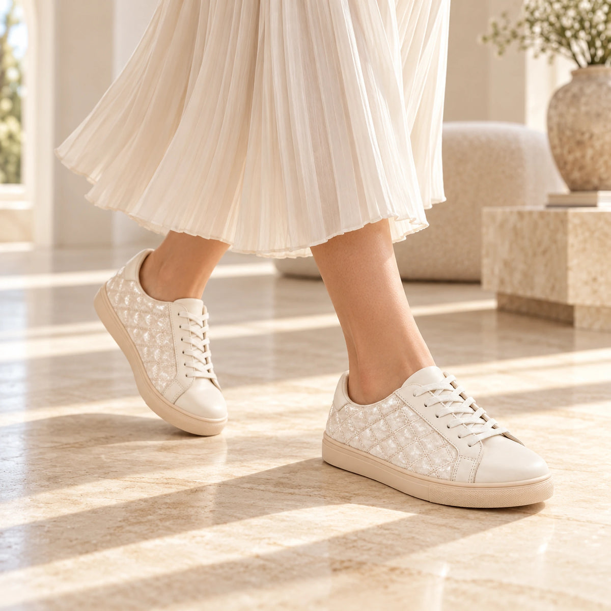 A person wearing white quilted lace-up sneakers with a pleated white skirt, standing on a polished beige marble floor in a bright, modern interior.