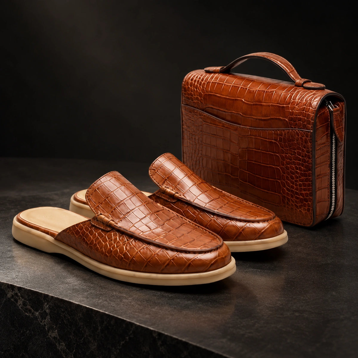 A pair of brown crocodile-embossed slip-on loafers with beige soles, displayed next to a matching brown crocodile-embossed zippered travel or vanity case on a dark stone surface against a dark background.