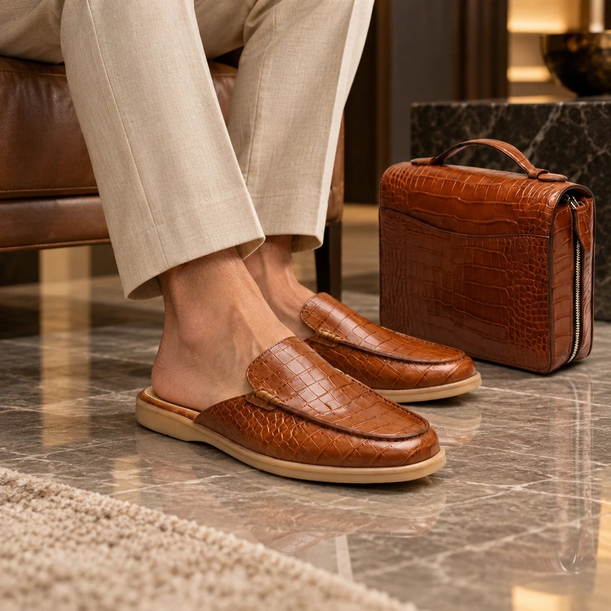 A person wearing brown crocodile-embossed slip-on loafers sits in a leather chair on a polished marble floor, with a matching brown crocodile-embossed zippered case beside them.