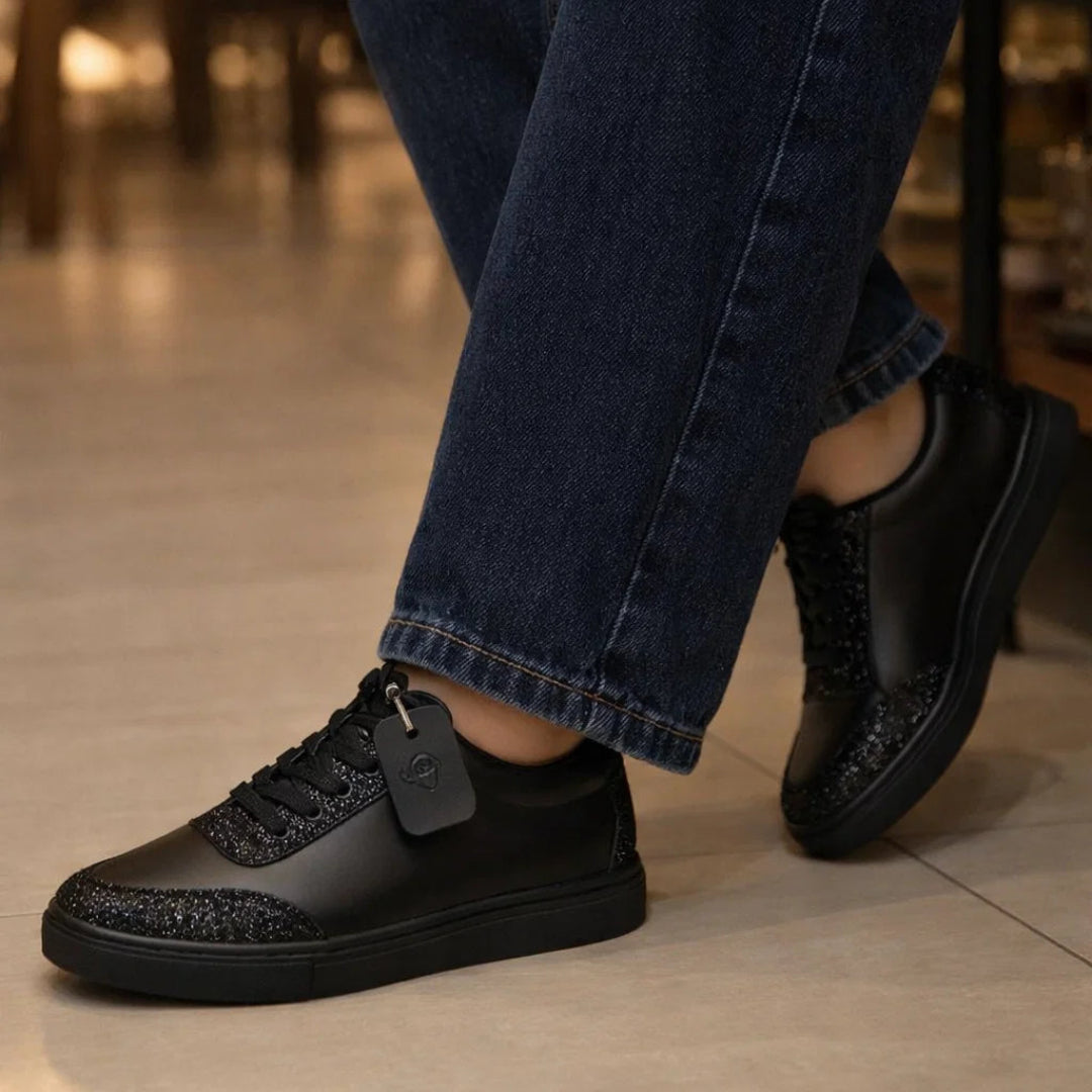 Black sneakers with glittery details worn with blue jeans on a blurred indoor background