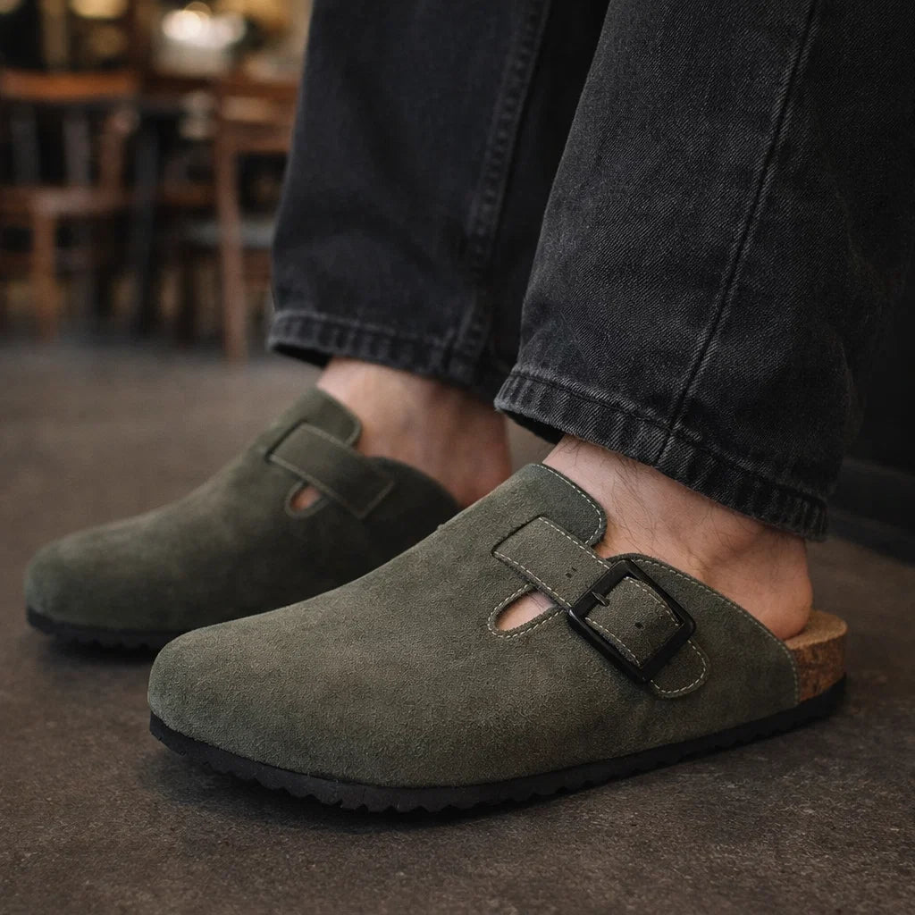 Green suede clogs worn with dark jeans on a blurred indoor background