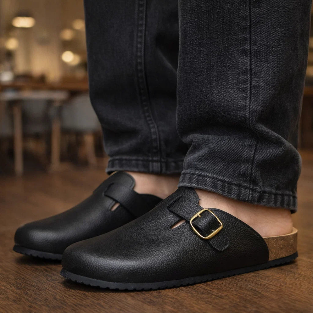 Black leather clogs with a gold buckle worn indoors on wooden flooring.
